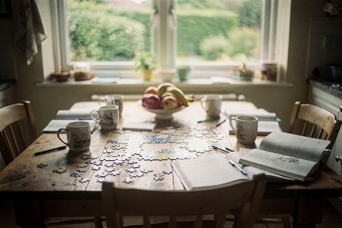 A kitchen table with mugs, a jigsaw and an open book by a sunny window